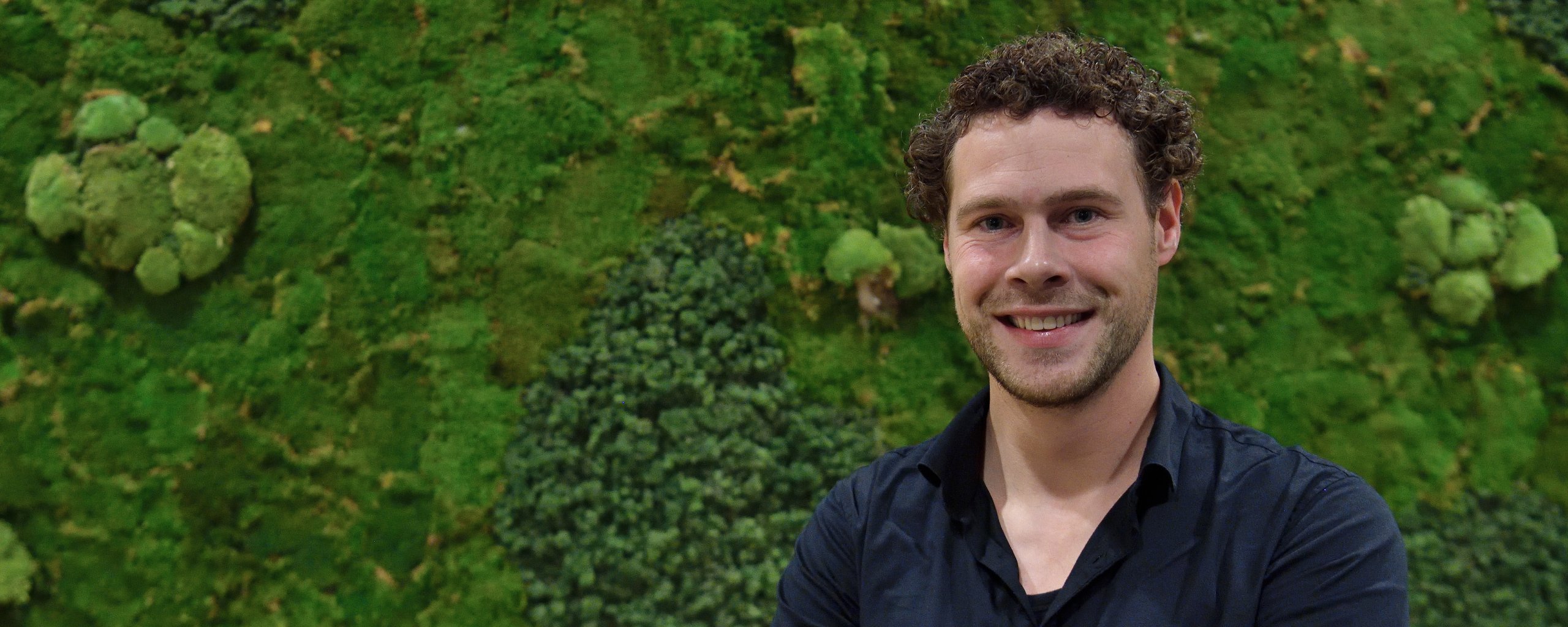 Een man staat met zijn armen over elkaar en een lach op het gezicht voor een wand vol groene mos.