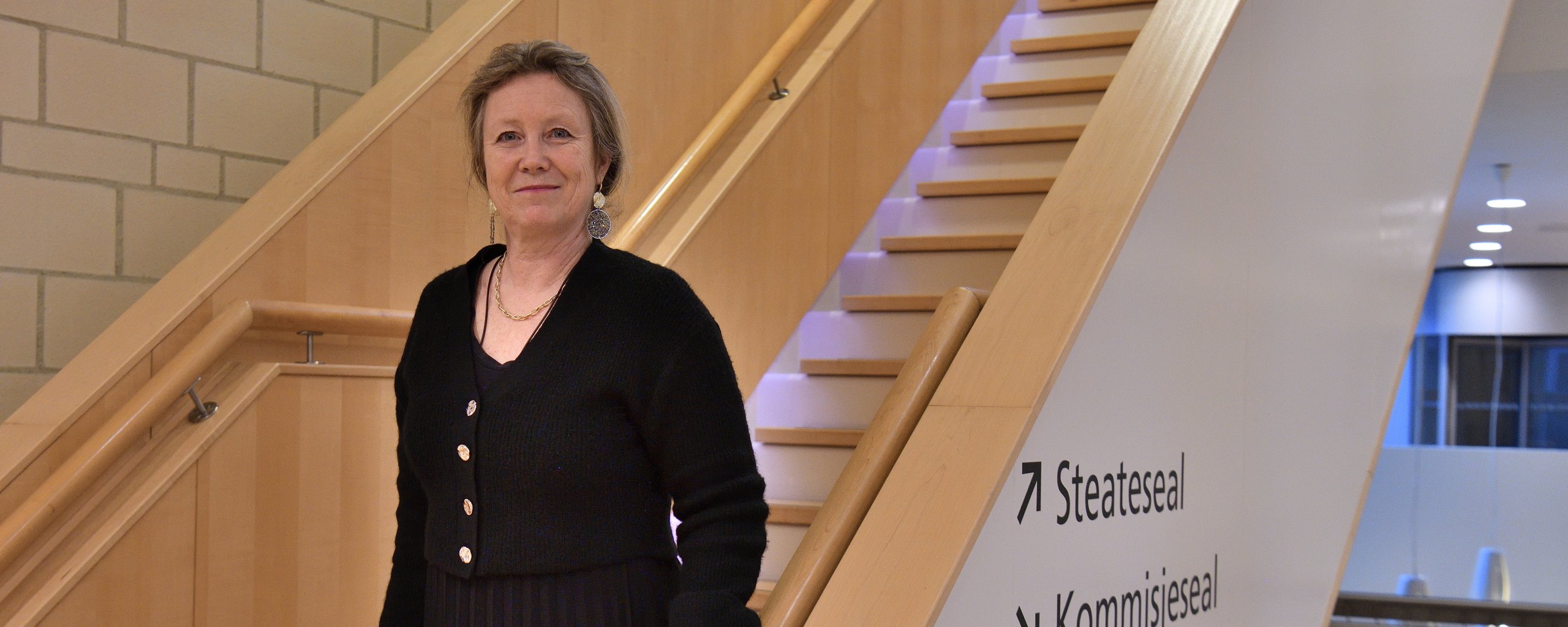 Een vrouw in zwart gekleed staat op een trap met haar hand op de leuning.