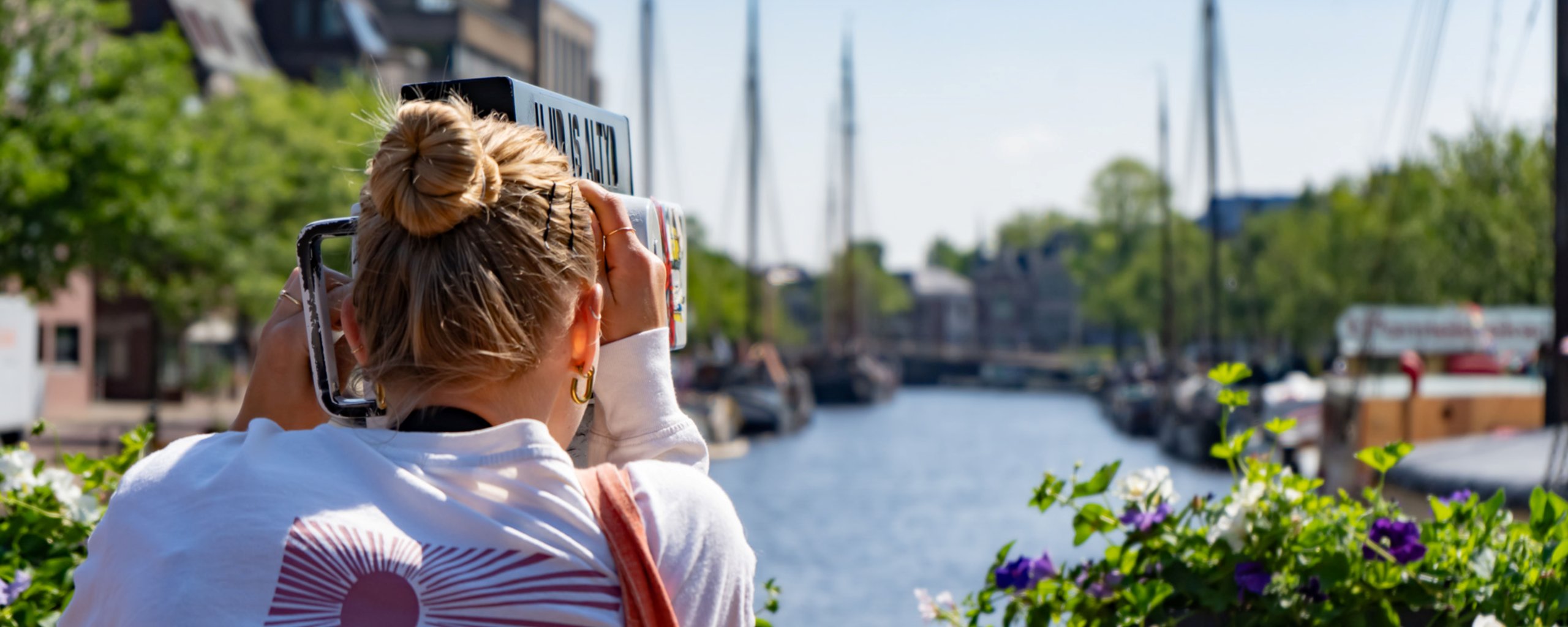 Een vrouw met een knotje in het haar kijkt door een verrekijker over het water waar allemaal bootjes liggen.