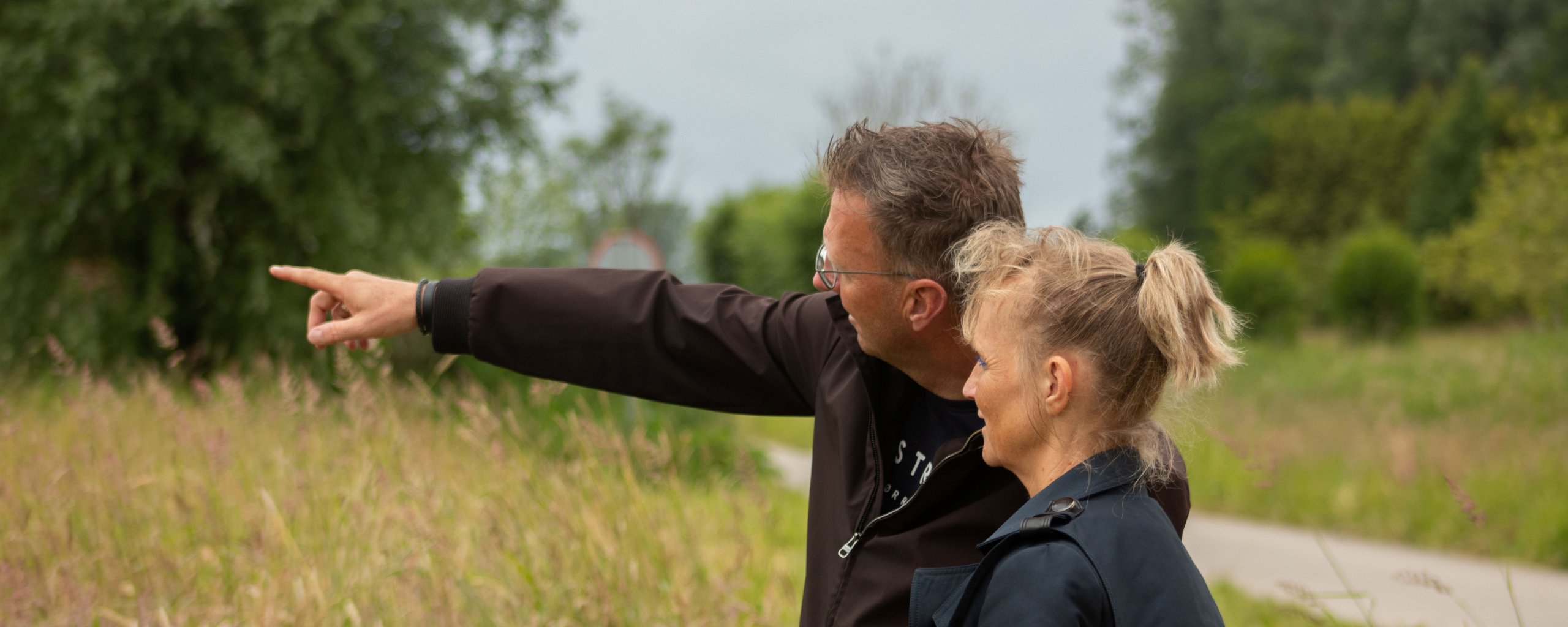 Een man en een vrouw staan naast elkaar in een bosrijke omgeving. de man wijst ergens naar en de vrouw kijkt die kant ook op.