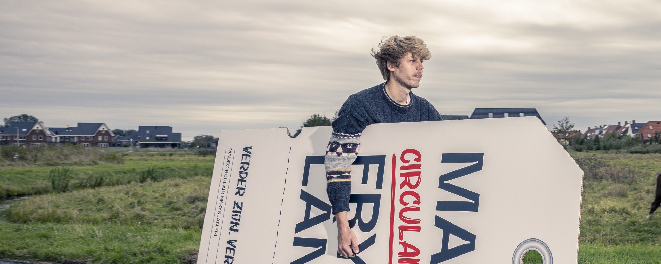 Jongeman op een fiets met een enorm groot label onder zijn arm waar op staat: Made in circulair Fryslân.