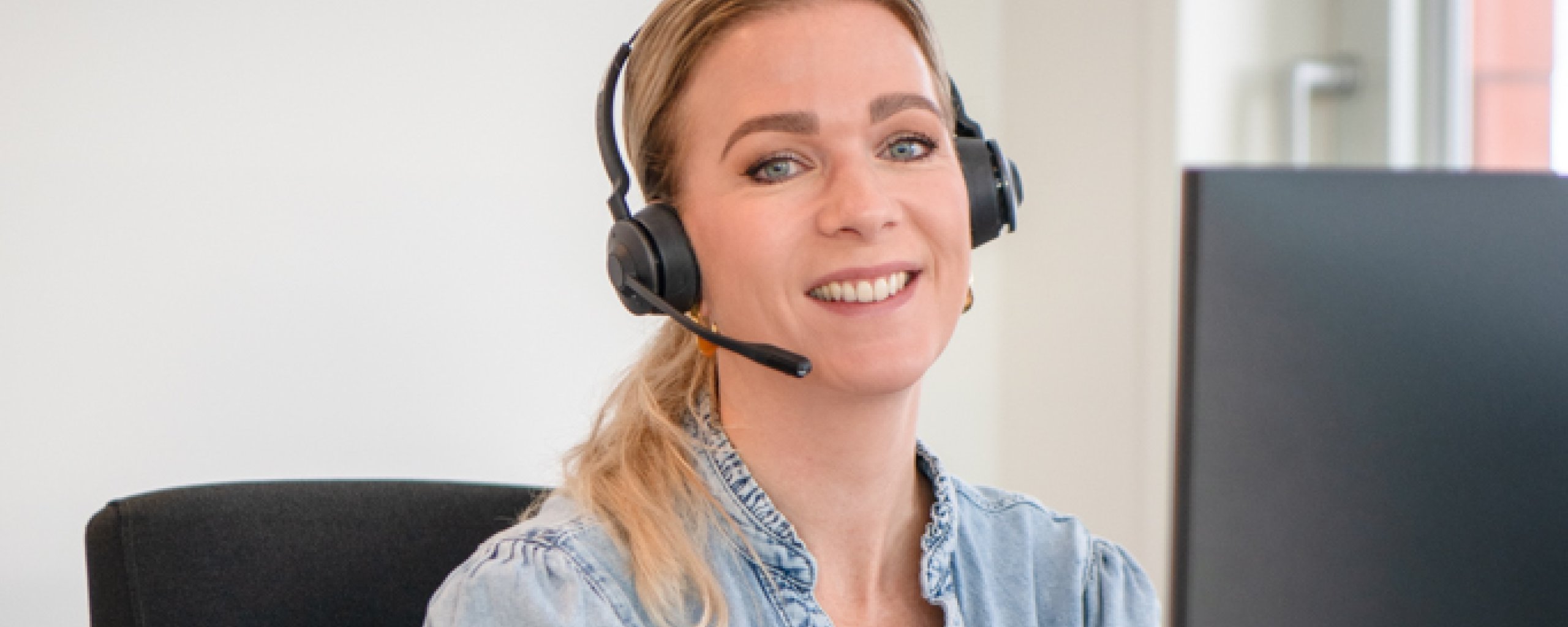 Een lachende vrouw met een headset op zit achter een computer.