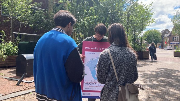 Drie personen staan in een kring te kijken naar een groot bord met een kaart daarop. Ze staan midden in een woonwijk.
