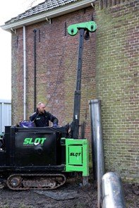 Een man is met een machine bezig om een paal in de grond te heien aan de buitenmuur.