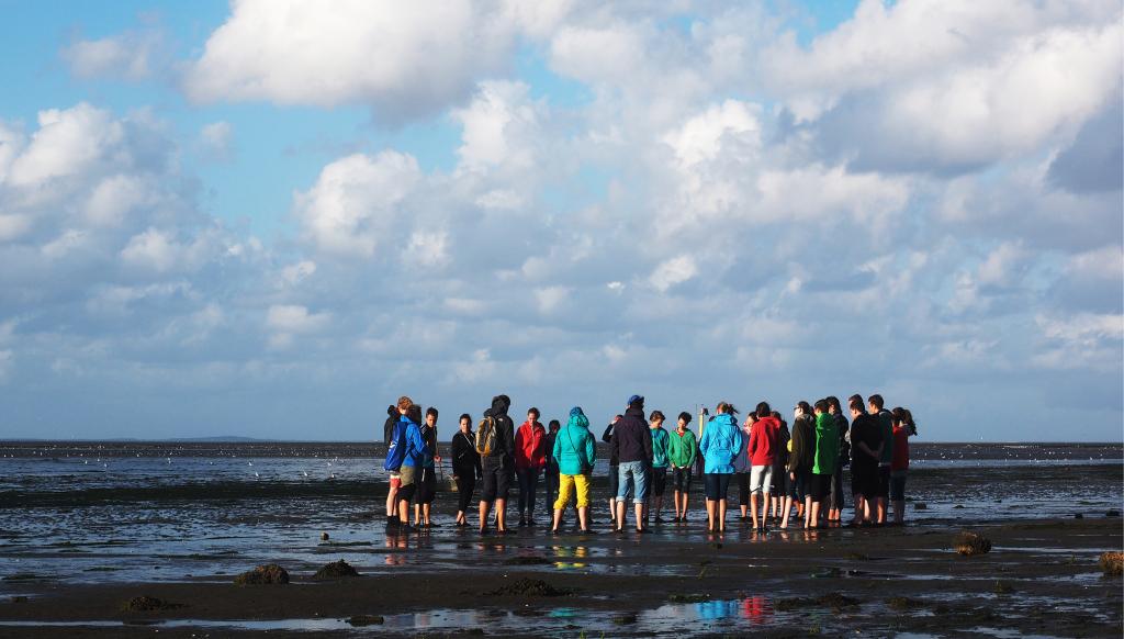 Een grote groep mensen staan in het natte zand met korte broeken en gekleurde regenjassen aan