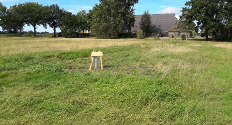 midden in een weiland, omzoomd door bomen, staan 3 kleine paaltjes met een bordje er op.