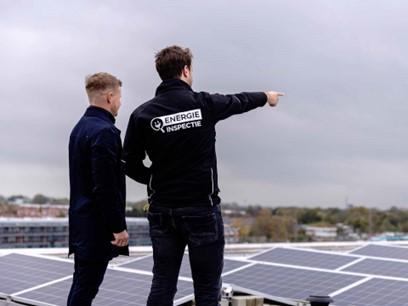 Twee personen staan uit te kijken op een zonnepanelenpark. De rechter persoon heeft een jasje van Energie Inspectie aan en wijst naar de zonnepanelen