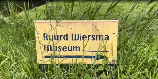 Een geel bord in het gras met blauwe letters en een pijl naar rechts wijzend. Er staat Ruurd Wiersma Museum op.