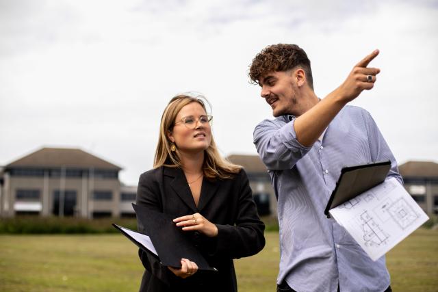 Een man en een vrouw staan in een weiland met een huis op de achtergrond. Beiden hebben een map met papieren in de hand en de man wijst met zijn vinger naar rechts.