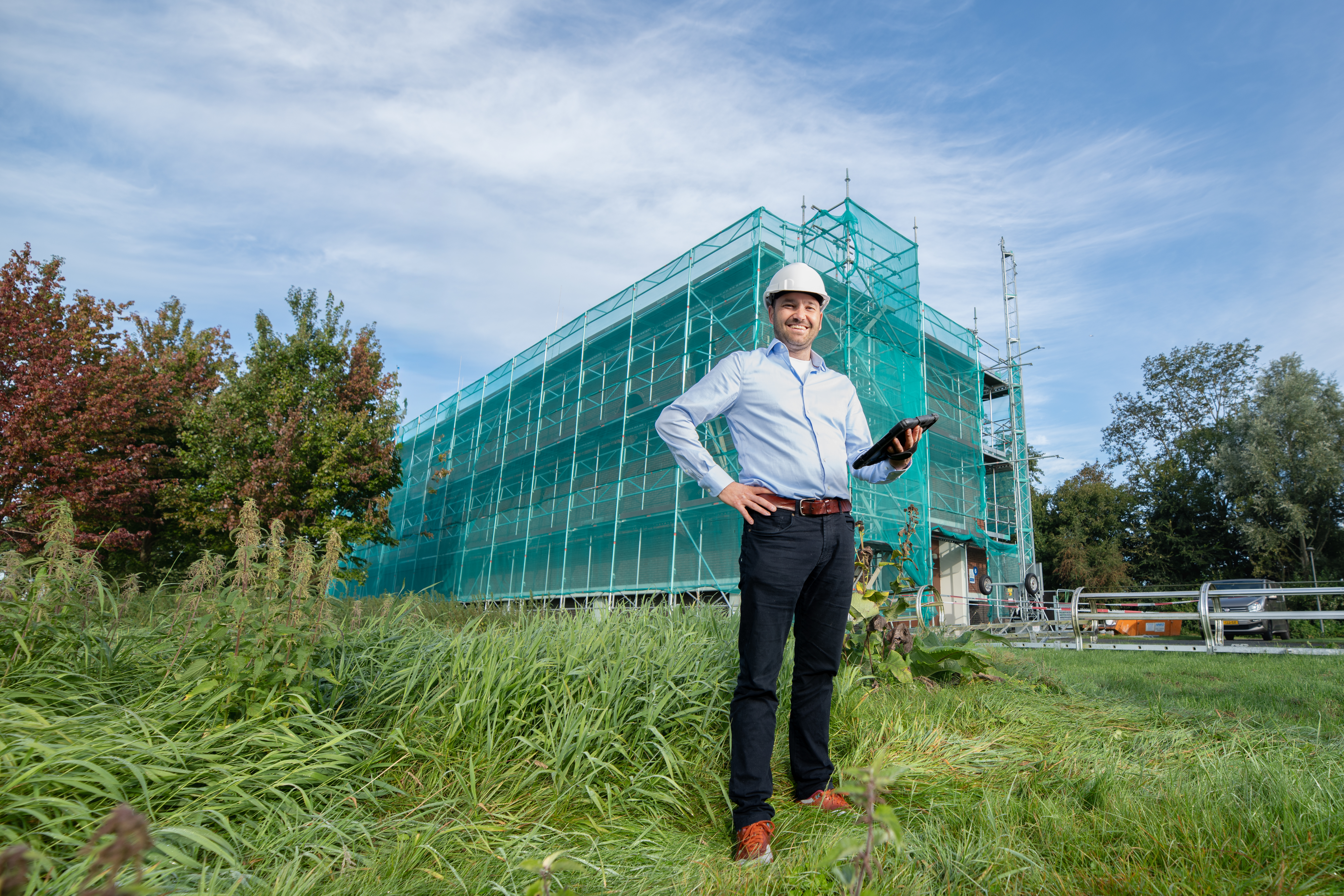 Een man met bouwhelm staat lachend voor een gebouw dat in de steigers staat.