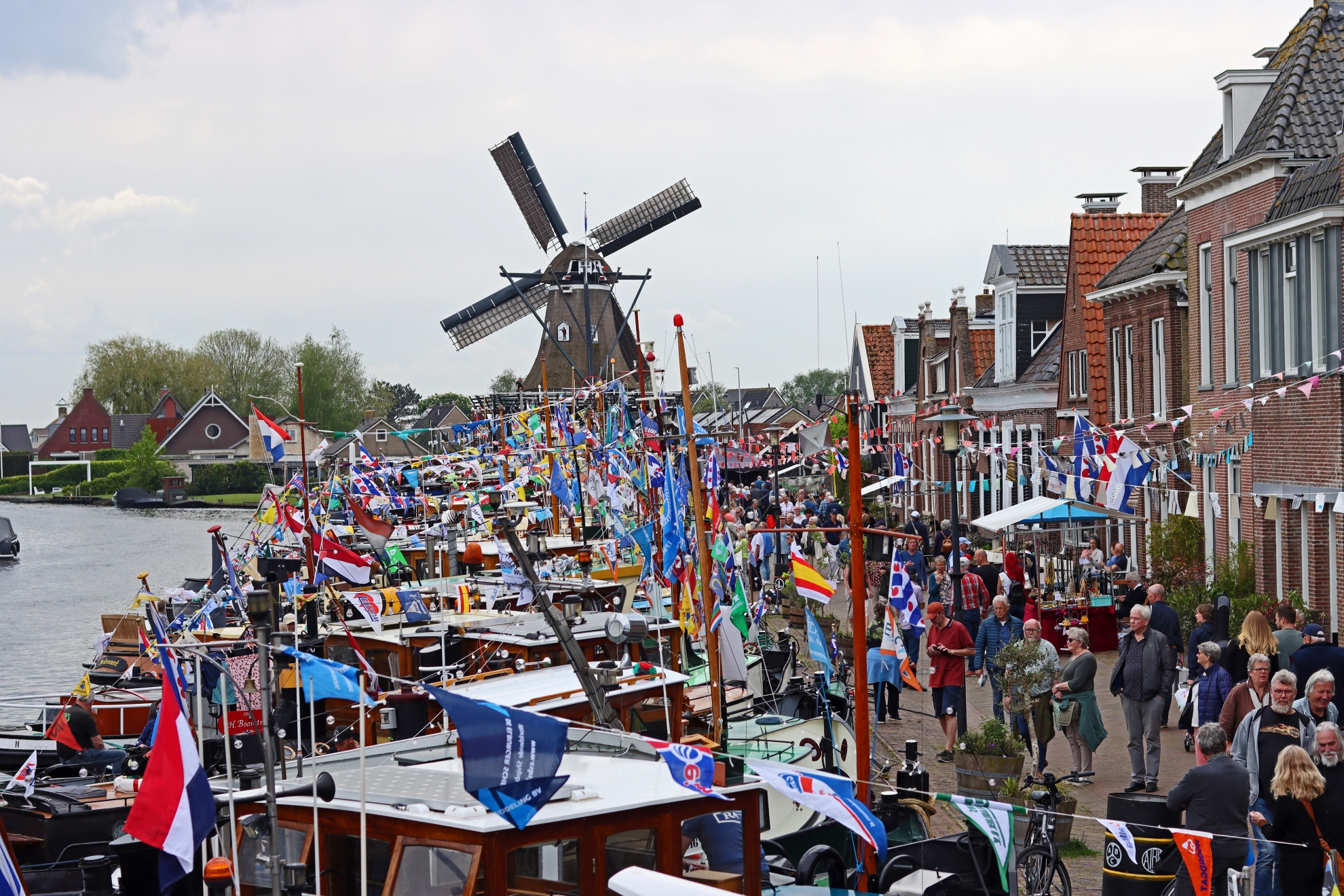 Een molen op de achtergrond en op de voorgrond allemaal boten die naast elkaar liggen. Er is allemaal volk op de been en er hangen overal vlaggetjes.