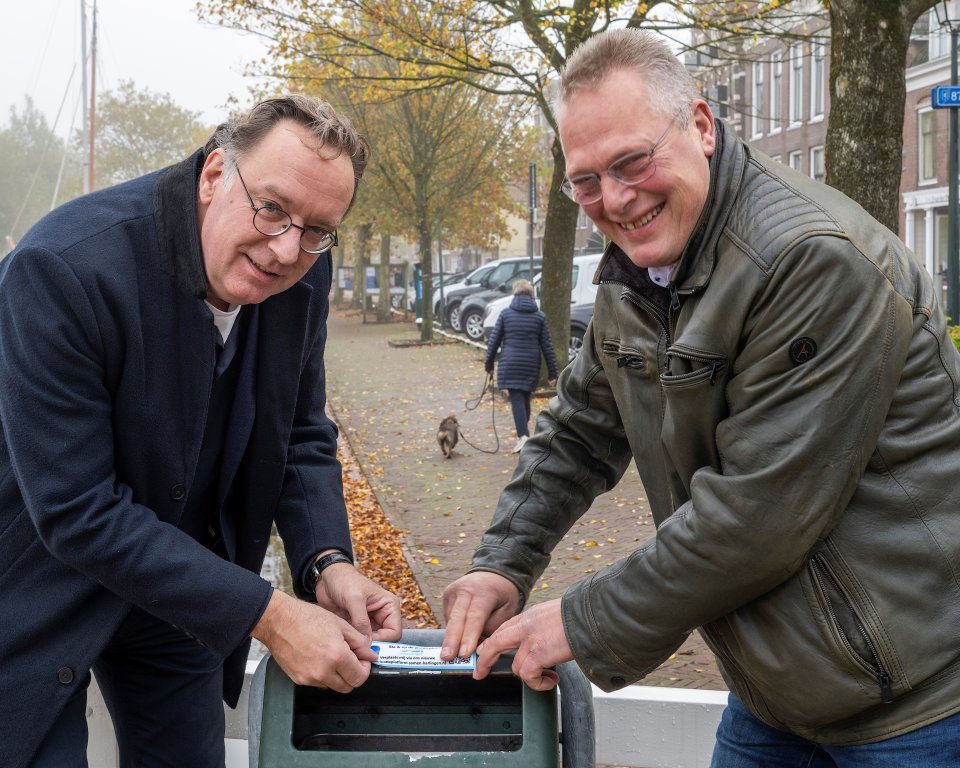 Wethouders Paul Schoute en Hendrik Sijtsma plakken een sticker een een afvalbak. 