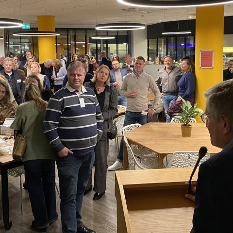 De wethouder staat achter een spreekzuil en spreekt een groep aanwezigen toe.