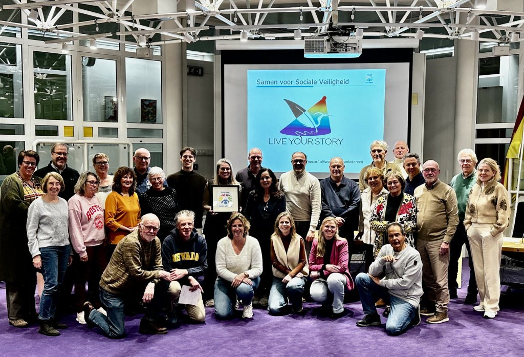 Deelnemers aan de bijeenkomst Samen voor Sociale Veiligheid samen op een groepsfoto. Op een groot scherm staat 'Samen voor Sociale Veiligheid, Live your story'.