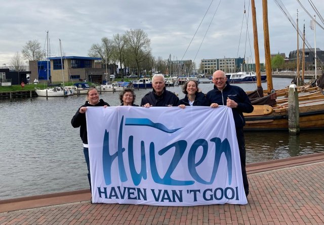 Vijf havenmeesters houden laggend een vlag omhoog met daarop de tekst 'Huizen, haven van het Gooi'.