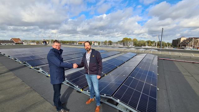 Wethouder Bert Rebel en bestuurslid Paul Schouten staan naast de zonnepanelen, bovenop het dak van de loods op het haventerrein van ’t Huizerhoofd.