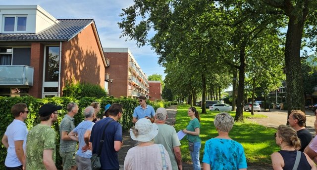 Een groep mensen staan in een cirkel naar een persoon te luisteren, midden op een groestrook in de wijk