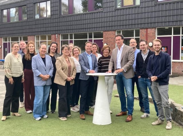 De wethouder staat samen met een groep mensen achter een tafel, voor een gebouw