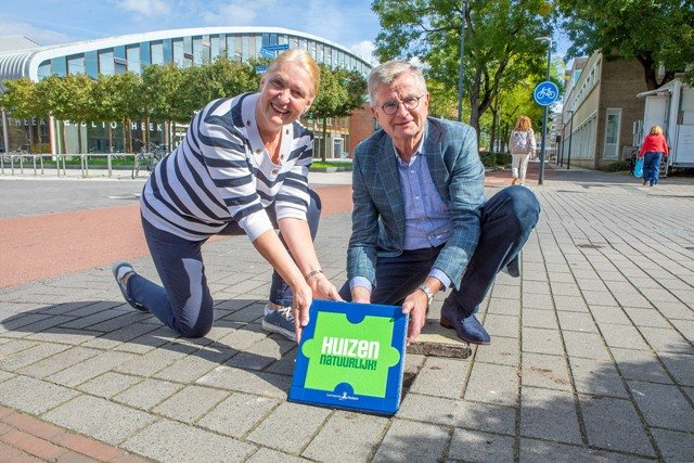 Wethouders Bert Rebel en Karin van Werven staan vlakbij het gemeentehuis en houden een stoeptegel vast waarop het logo van de duurzaamheidscampagne staat