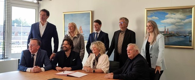 De collegeleden en andere betrokken personen zitten aan een vergadertafel met daarop een ondertekend document