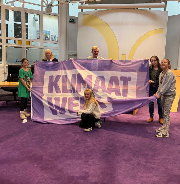 Wethouders Bert Rebel en Karin van Werven staan samen met 4 kinderen in de raadzaal van het gemeentehuis en houden een grote vlag omhoog, met daarop de tekst: Klimaatweek.