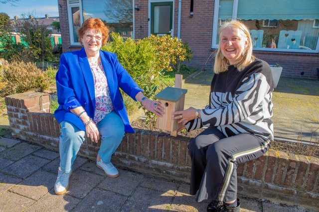 Wethouder Van Werven en bewoonster Tineke zitten in de zonnige voortuin en de wethouder overhandigt een vogelhuisje.
