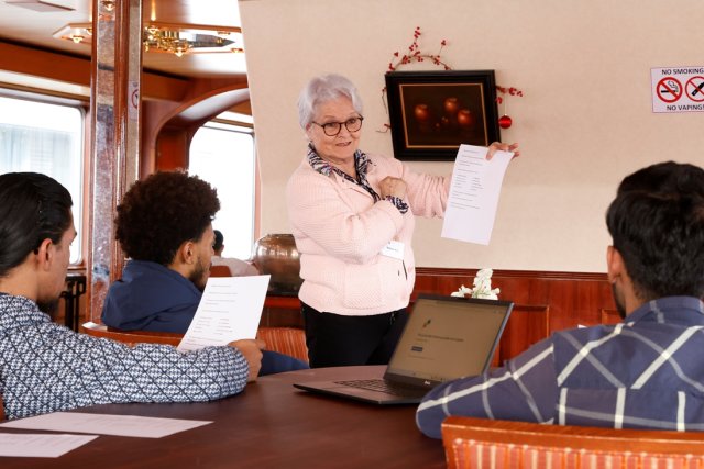 Magda staat in een opvangschip en houdt een A4-papier omhoog en kijkt 3 leerlingen aan