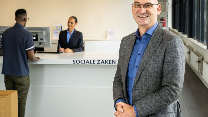 Frans van der Meij staat bij de balie van Sociale Zaken. Op de achtergrond wordt iemand daar geholpen door een medewerker.