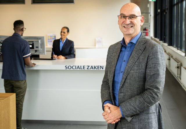 Frans van der Meij staat bij de balie van Sociale Zaken. Op de achtergrond wordt iemand daar geholpen door een medewerker.