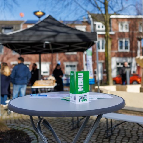 Een foto van een statafel met daarop een folder met de tekst 'menu'. Op de achtergrond is een kleine overkapping met tentdoek zichtbaar, waar mensen onder staan. 