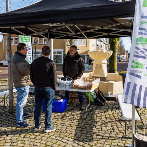 Wethouder van Drunen staat onder een overkapping bij de Sjpringbron te praten met twee anderen. Tevens is de vlag van 't Kirchroa va Mörje zichtbaar. 
