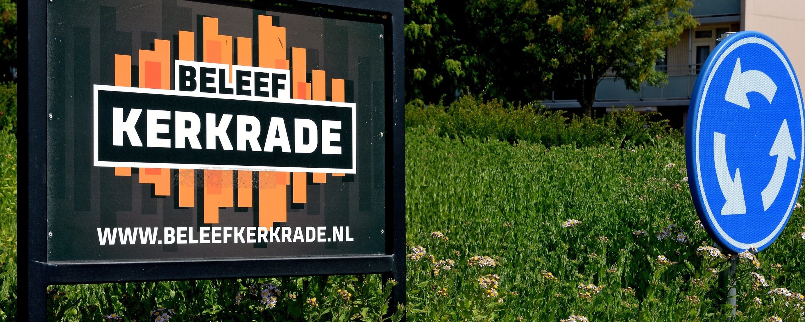 Links op de foto een reclamebord met het Beleef Kerkrade logo, rechts het blauwe rotondebord. De rotonde staat vol groen. Op de achtergrond groene bomen en een flat.