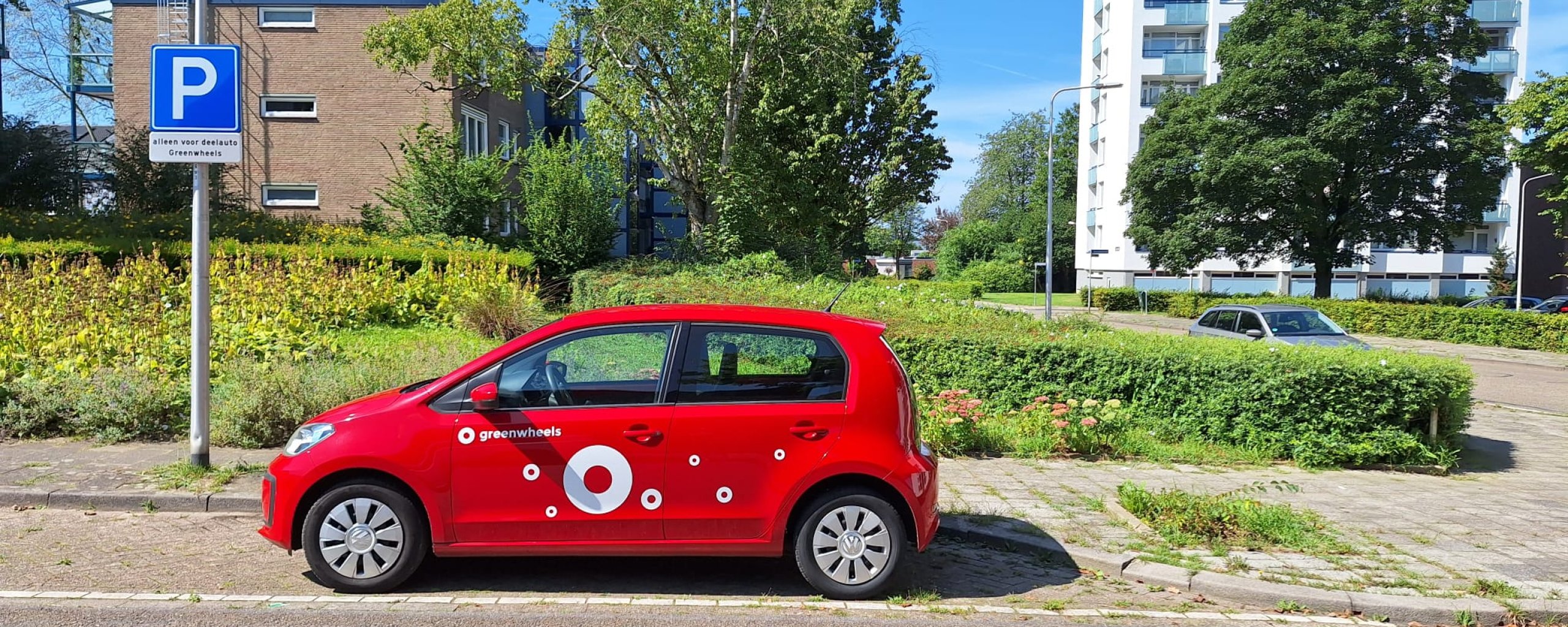 Een rode Volkswagen UP staat op een deelauto parkeerplaats aan de Kremerstraat. Op de achtergrond zie je gebouwen. 