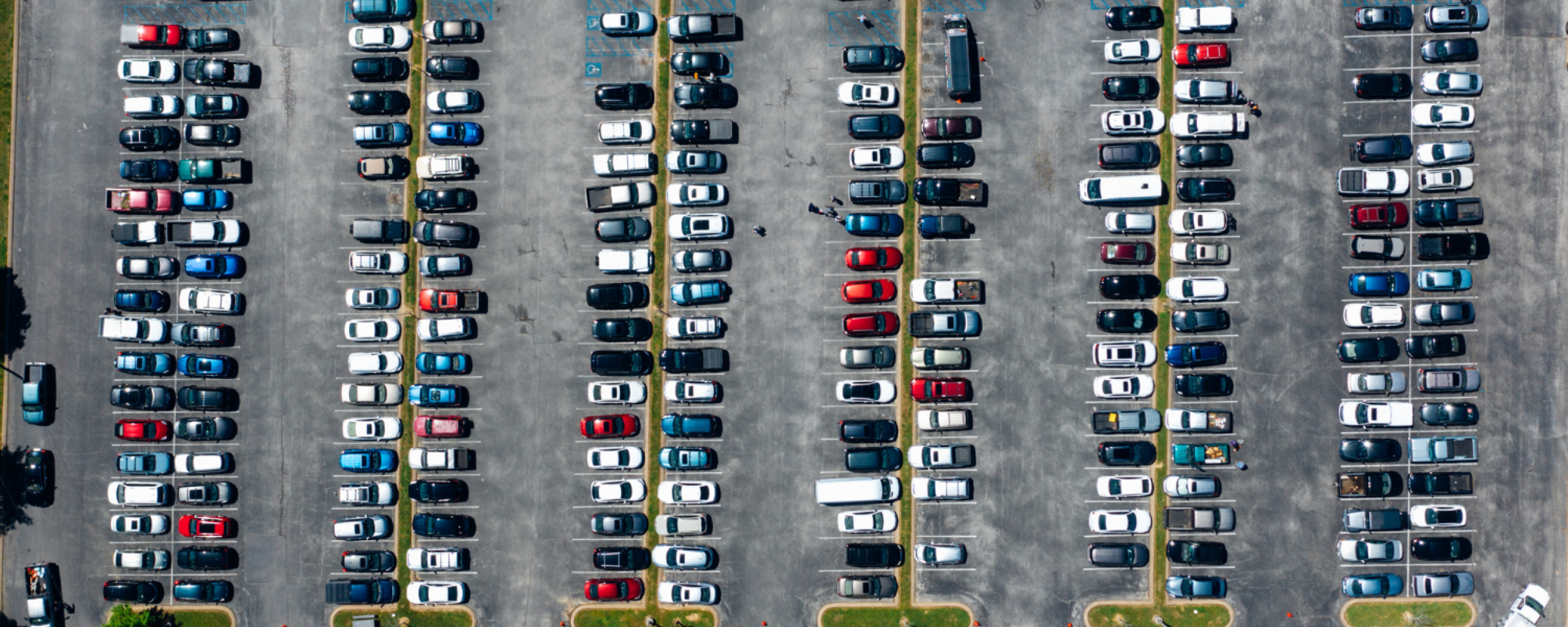 Een kleurrijk bovenaanzicht van een parkeerterrein gevuld met auto's. Op en rond het terrein liggen grasstroken. 
