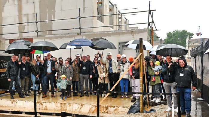 Een groep mensen voor de huizen in aanbouw op de Gracht. Het regent en meerdere mensen houden een paraplu in de lucht. Op de voorgrond een kindje met een bouwhelm op en een troffel in zijn hand. 
