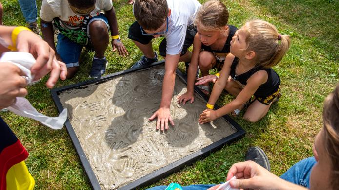 Een aantal kinderen laat een handafdruk achter in beton. De plaat met beton ligt op het gras.