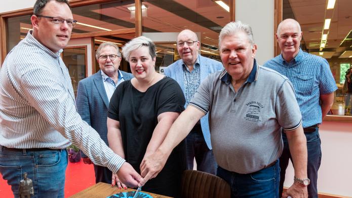 Drie personen snijden een blauwe taart aan waarop staat 'ruilwinkel Kerkrade' Wethouder Weiermans kijkt op afstand met nog twee personen toe. Alle personen kijken in de camera. 