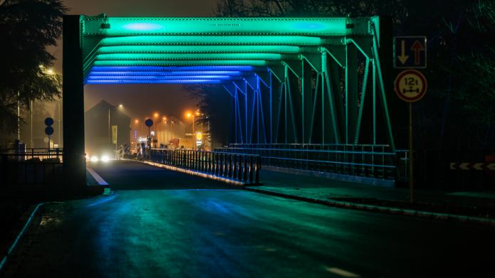De verlichte spoorbrug in het donker. De brug licht blauw en groen op. Op de achtergrond de rotonde  en de Lidl. 