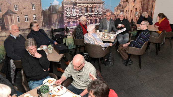 Mensen aan tafeltjes tijdenss de Kaffieëklatsj mit d’r Jo op de achtergrond een foto van het Raadhuis als wand. 