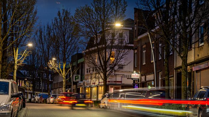 Twee straatlantaarns met LED verlichting  die branden in het donker in een straat met huizen, er komen auto's voorbij die met lange sluitertijd gefotografeerd zijn