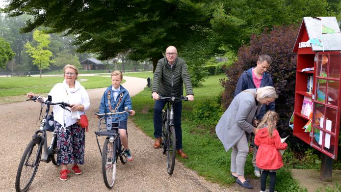 Van links naar rechts organisator Marleen Fischer, kinderburgemeester Hidde Sieler en wethouder Jo Schlangen op de fiets bij een minibieb waar een gezin een boek uitzoekt
