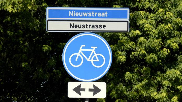 Straatnaambord Nieuwstraat in Nederlands en Duits tegen een achtergrond van bomen met daaronder een bord fietspas in beide richtingen. 