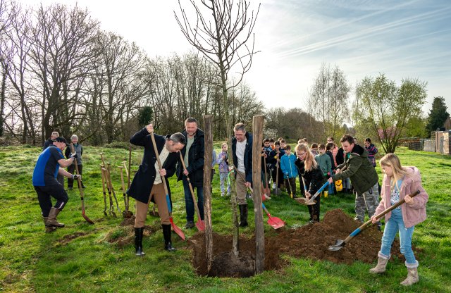 Wethouder Geers plant met kinderen een jonge kastanjeboom. Ze scheppen het gat dicht waarin de boom is neergezet. 