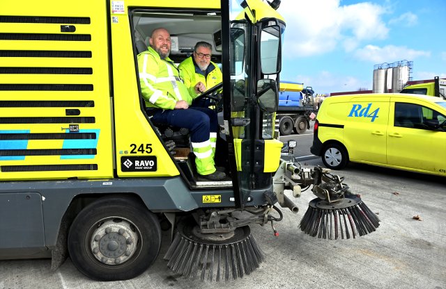 Wethouder Van Drunen poseert samen met chauffeur Mark Smeets in de stuurcabine van een gele veegwagen van Rd4. Ze dragen allebei gele veiligheidskleding.