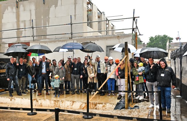 Een groep mensen voor de huizen in aanbouw op de Gracht. Het regent en meerdere mensen houden een paraplu in de lucht. Op de voorgrond een kindje met een bouwhelm op en een troffel in zijn hand. 