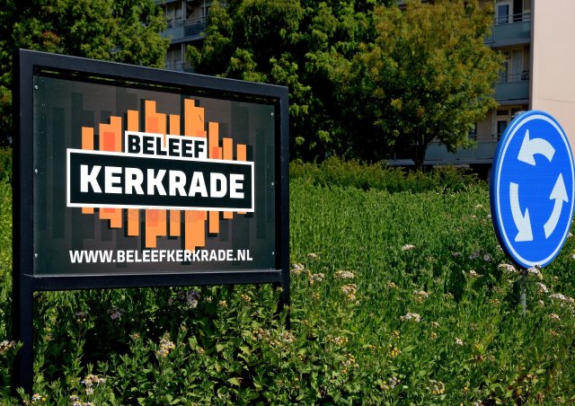 Links op de foto een reclamebord met het Beleef Kerkrade logo, rechts het blauwe rotondebord. De rotonde staat vol groen. Op de achtergrond groene bomen en een flat.