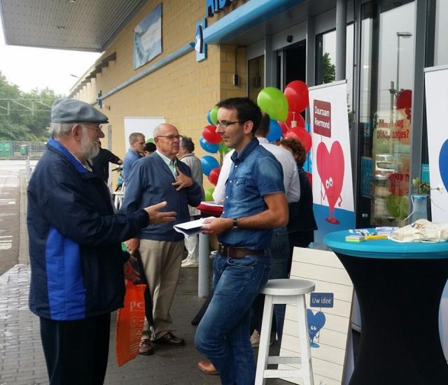 Mensen praten met elkaar op een infostand onder de overkapping van een gebouw. Er staan roll-up banners, er hangen gekleurde ballonnen, er staan statafels en krukken