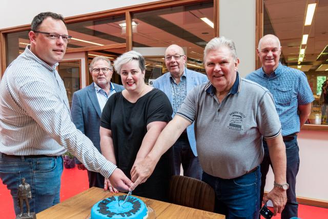 Drie personen snijden een blauwe taart aan waarop staat 'ruilwinkel Kerkrade' Wethouder Weiermans kijkt op afstand met nog twee personen toe. Alle personen kijken in de camera. 