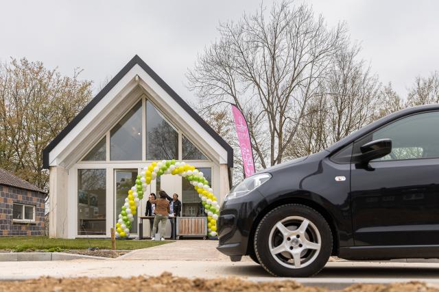 Een zwarte auto staat voor één van de SUPERLOCAL proefwoningen, voor de woning staat een tafel waar mensen kunnen tekenen, boven de tafel een haag van ballonnen. 