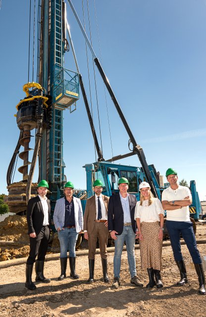 Een groep mensen met bouwhelmen op een bouwterrein en op de achtergrond een heimachine